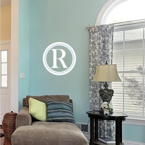 Könnte beinhalten: Ein Wohnzimmer mit einer Chaiselongue, Kissen und einem Beistelltisch. Die Wand ist hellblau gestrichen und zeigt ein weißes "R" in einem Kreis. Auf dem Beistelltisch steht eine Lampe neben einem Fenster mit gemusterten Vorhängen.