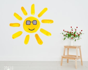 May include: A yellow sun with a smiling face and sunglasses is painted on a white wall. A small wooden step stool is in the foreground with a vase of red and white flowers on top.
