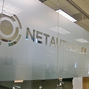 以下が含まれることがあります： すりガラスのオフィス窓。ロゴと「NETAUTHORITY」のテキストがサンセリフ体で表示されています。ロゴは、一連の同心円状の形状を持つ円形のデザインです。ガラスを通してオフィスの内部が見えます。