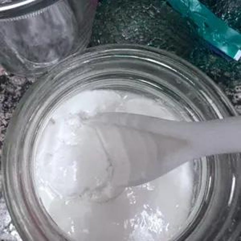 Puede incluir: Un tarro de vidrio transparente lleno de una sustancia blanca espesa. Una cuchara blanca est&aacute; parcialmente sumergida, removiendo la sustancia. El tarro est&aacute; abierto y el contenido parece suave y cremoso. El fondo est&aacute; ligeramente borroso.