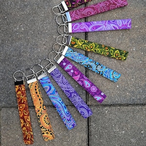 May include: A collection of colorful fabric wrist keychains with silver key rings. The keychains feature various paisley and floral patterns in shades of purple, blue, green, orange, and brown. The keychains are arranged on a gray stone surface.