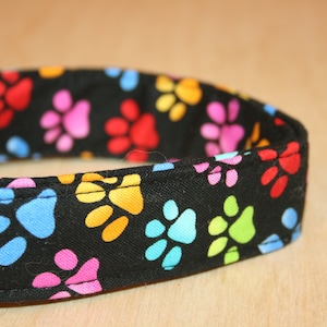 May include: A black dog collar with a colorful paw print pattern. The collar is made of fabric and has a black plastic buckle.