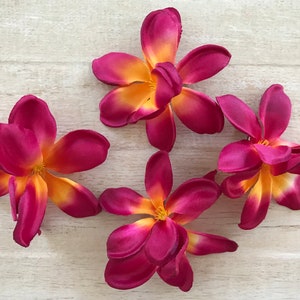 May include: Four artificial plumeria flowers with pink petals and yellow centers. The flowers are arranged on a light wood surface.