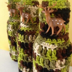 May include: Three glass bottles with green tops are covered in crocheted bottle cozies. The cozies are a camouflage pattern of green, brown, and beige yarn. Each bottle cozy has a small plastic deer figurine attached.