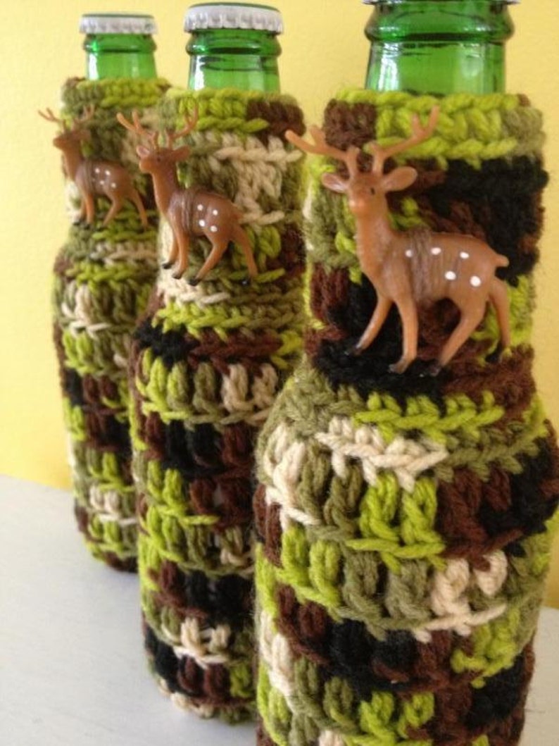 May include: Three glass bottles with green tops are covered in crocheted bottle cozies. The cozies are a camouflage pattern of green, brown, and beige yarn. Each bottle cozy has a small plastic deer figurine attached.
