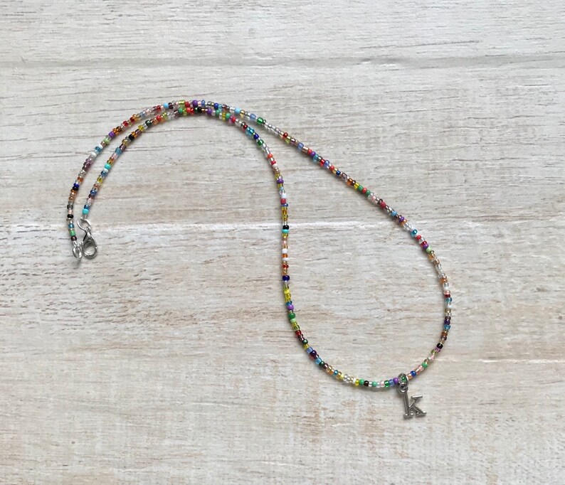 May include: A colorful beaded necklace with a silver letter K charm. The necklace is made of small, multi-colored beads and has a silver clasp.