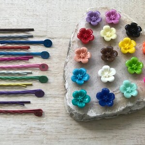 May include: Assortment of colorful flower-shaped hair clips with a variety of colors including pink, red, orange, yellow, green, blue, purple, and brown. The clips are arranged on a light brown rock-like surface.