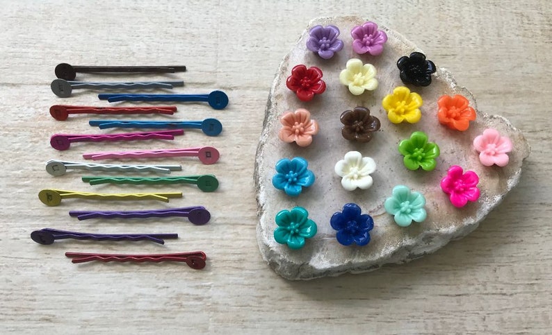 May include: Assortment of colorful flower-shaped hair clips with a variety of colors including pink, red, orange, yellow, green, blue, purple, and brown. The clips are arranged on a light brown rock-like surface.