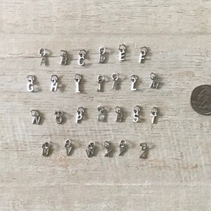 May include: A set of 26 silver letter charms, each charm features a different letter of the alphabet. The charms are arranged in alphabetical order on a wooden surface.