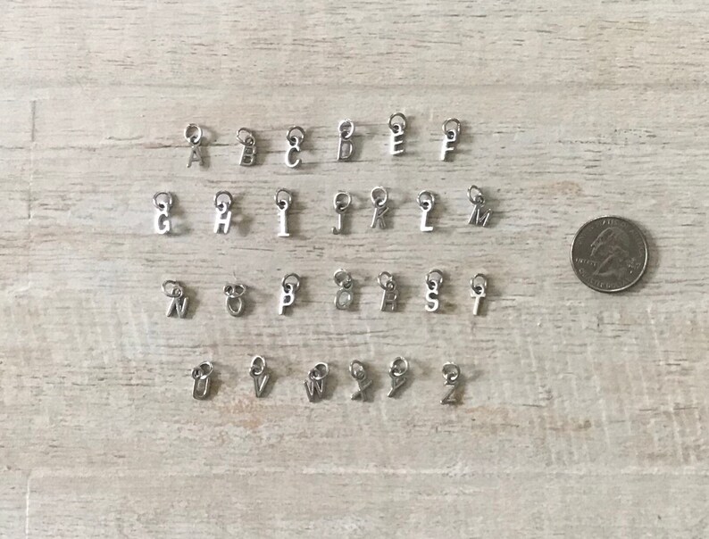 May include: A set of 26 silver letter charms, each charm features a different letter of the alphabet. The charms are arranged in alphabetical order on a wooden surface.