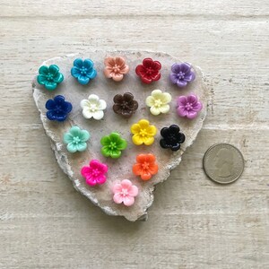 May include: Assortment of 18 colorful flower-shaped buttons, arranged in a heart shape on a gray stone surface. A US quarter is included for size reference.