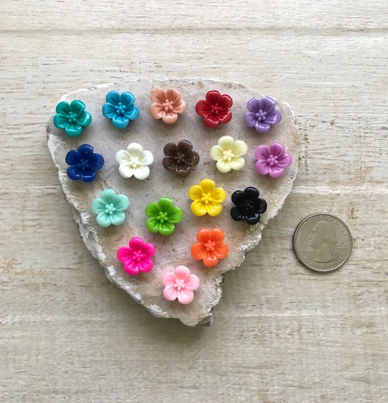 May include: Assortment of 18 colorful flower-shaped buttons, arranged in a heart shape on a gray stone surface. A US quarter is included for size reference.