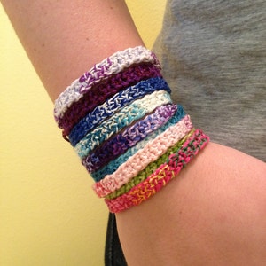May include: A stack of colorful crocheted bracelets in shades of pink, green, blue, purple, and white. The bracelets are worn on a person's wrist.