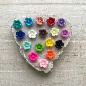 May include: Assortment of 16 colorful flower-shaped buttons in various colors, including red, blue, green, yellow, pink, orange, and white. The buttons are arranged in a heart shape on a light brown wooden surface.