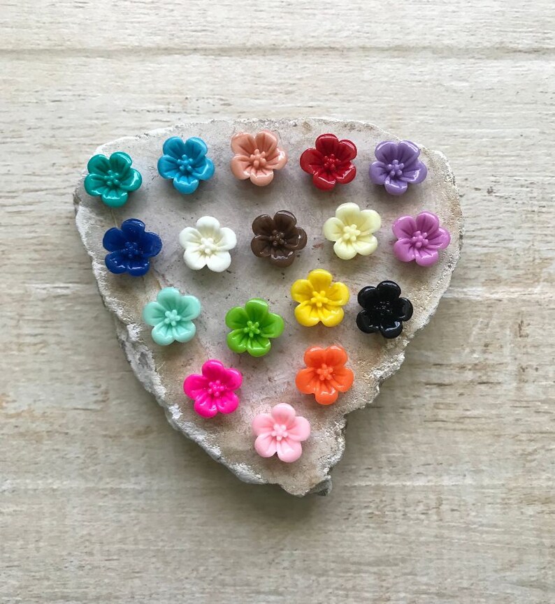 May include: Assortment of 16 colorful flower-shaped buttons in various colors, including red, blue, green, yellow, pink, orange, and white. The buttons are arranged in a heart shape on a light brown wooden surface.