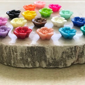 May include: Assortment of 16 colorful flower-shaped buttons in various colors, including pink, purple, red, orange, yellow, green, blue, and brown. The buttons are arranged on a wooden surface.