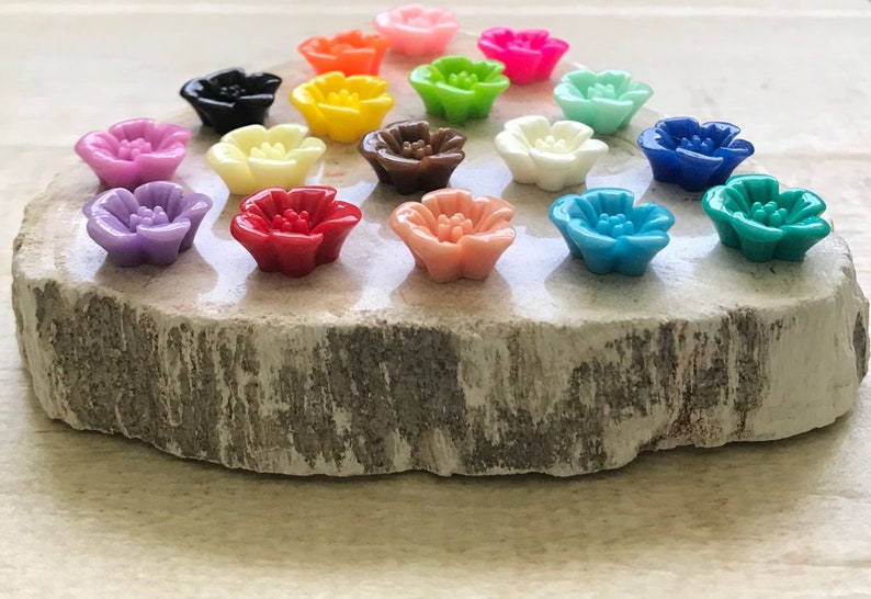 May include: Assortment of 16 colorful flower-shaped buttons in various colors, including pink, purple, red, orange, yellow, green, blue, and brown. The buttons are arranged on a wooden surface.