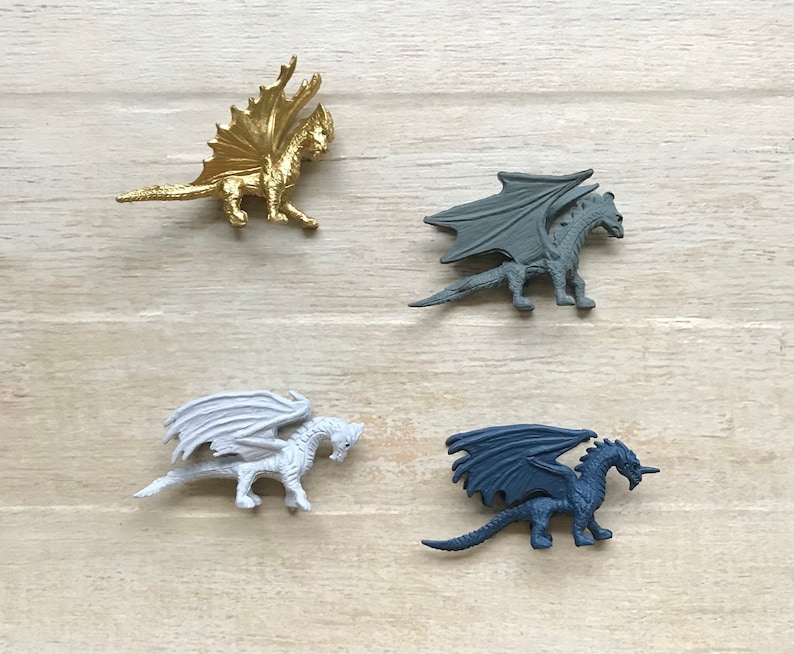 May include: Four miniature dragon figurines in different colors: gold, gray, white, and dark blue. The dragons are posed in a flying position with wings spread.