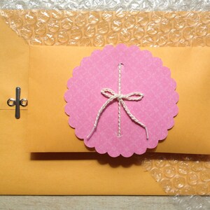 May include: A pink envelope with a scalloped edge and a white bow. The envelope is sealed with a silver clasp. The envelope is sitting on a sheet of bubble wrap.