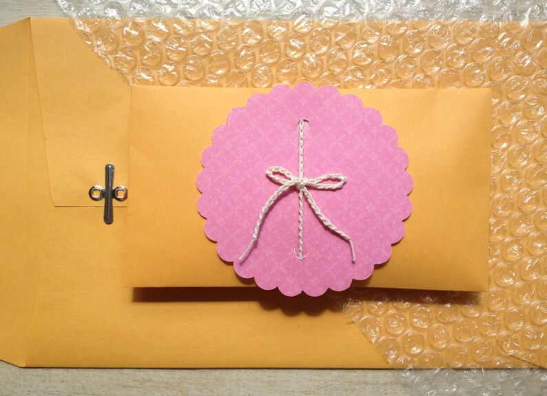 May include: A pink envelope with a scalloped edge and a white bow. The envelope is sealed with a silver clasp. The envelope is sitting on a sheet of bubble wrap.