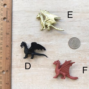 May include: Three plastic dragon figurines, one gold, one black, and one red. The dragons are posed in a standing position with their wings spread out. The figurines are arranged on a light brown wooden surface with a ruler and a coin in the background.
