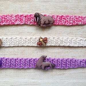 May include: Three crocheted bracelets with different colored yarn and buttons. The first bracelet is pink and white with a brown walrus button. The second bracelet is white with a brown bee button. The third bracelet is purple with a brown seal button.