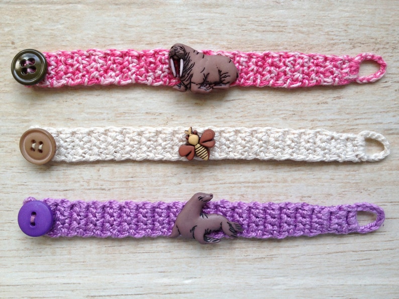 May include: Three crocheted bracelets with different colored yarn and buttons. The first bracelet is pink and white with a brown walrus button. The second bracelet is white with a brown bee button. The third bracelet is purple with a brown seal button.