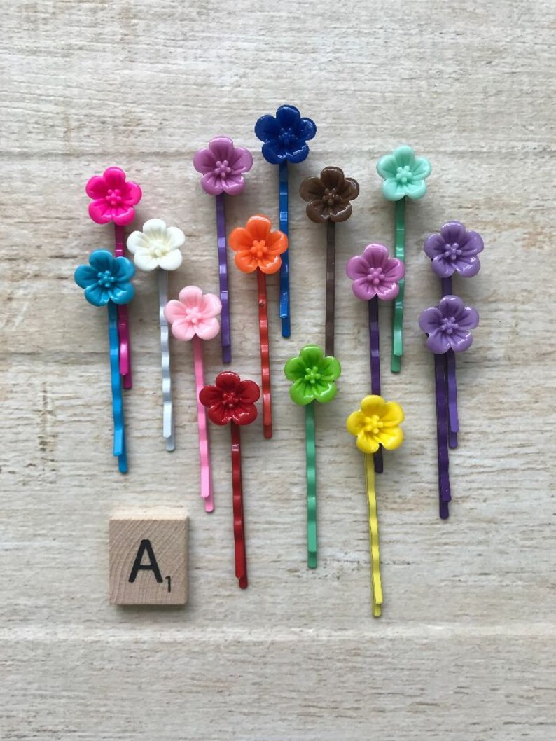 May include: Assortment of 14 colorful flower hair clips with a wavy metal bar. The clips are arranged on a wooden surface with a letter tile that reads "A1".