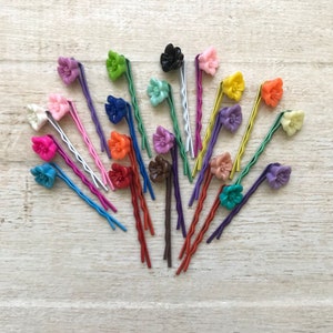 May include: A collection of 20 colorful flower hair clips with wavy metal pins. The clips are arranged in a fan shape on a light wood surface.