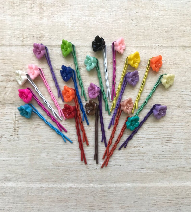 May include: A collection of 20 colorful flower hair clips with wavy metal pins. The clips are arranged in a fan shape on a light wood surface.