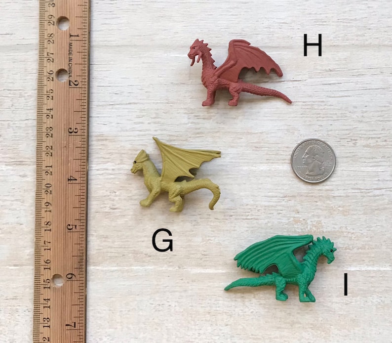 May include: Three plastic toy dragons, one red, one green, and one yellow, are arranged on a wooden surface with a ruler and a US quarter for scale. The dragons are all facing the right side of the image.