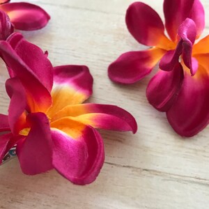 May include: Three burgundy and orange fabric plumeria flower hair clips. The flowers have a yellow center and are made of a soft, silky fabric.