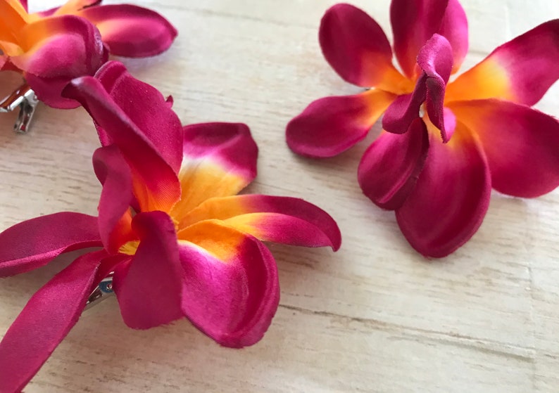 May include: Three burgundy and orange fabric plumeria flower hair clips. The flowers have a yellow center and are made of a soft, silky fabric.