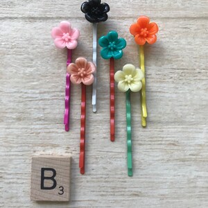 May include: Set of seven colorful flower hair clips with a black flower hair clip. The hair clips are on a wooden surface with a letter tile that says "B3".