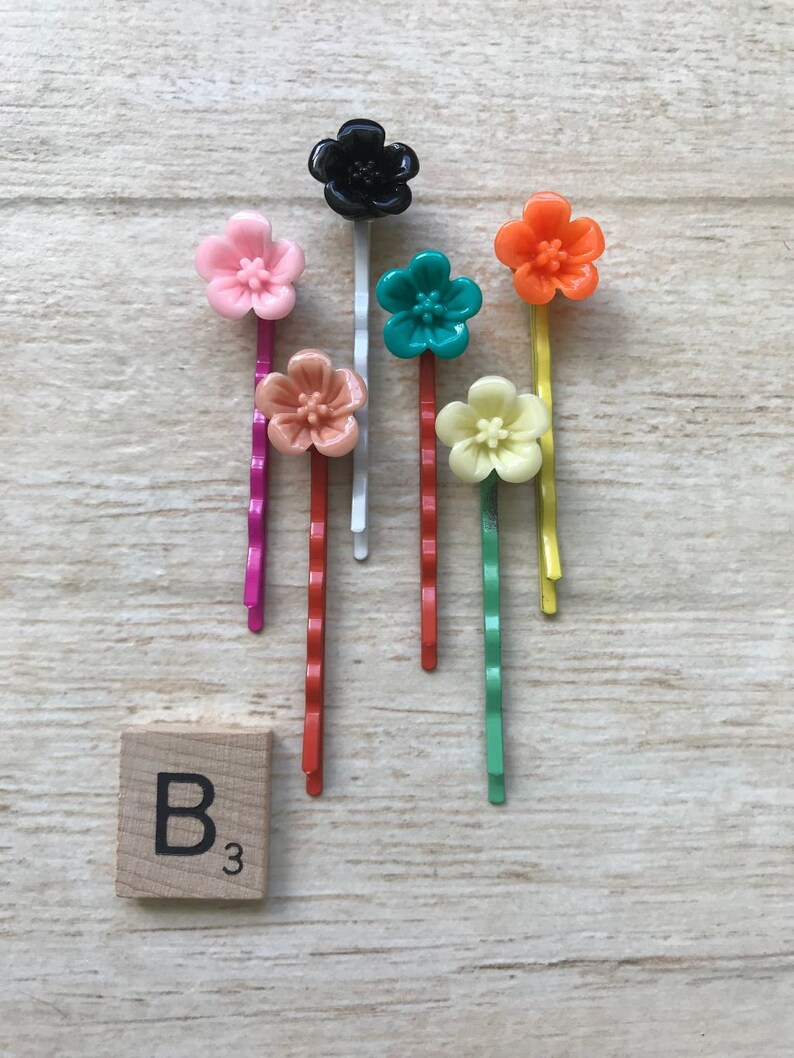 May include: Set of seven colorful flower hair clips with a black flower hair clip. The hair clips are on a wooden surface with a letter tile that says "B3".