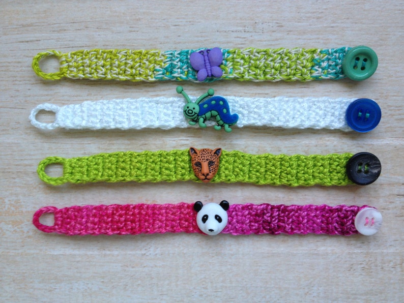 May include: Four crocheted bracelets with different colored buttons and charms. The bracelets are green, white, lime green, and pink. The charms include a butterfly, a caterpillar, a leopard, and a panda.