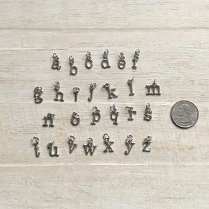 May include: A set of 26 silver letter charms, each charm is a letter of the alphabet. The charms are arranged in alphabetical order on a light wood surface.