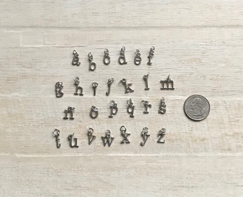 May include: A set of 26 silver letter charms, each charm is a letter of the alphabet. The charms are arranged in alphabetical order on a light wood surface.