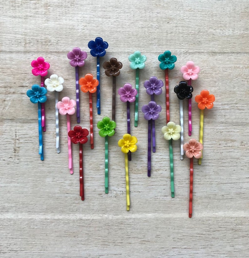 May include: A set of 18 colorful flower hair clips. The clips are made of plastic and have a variety of colors, including pink, blue, green, yellow, orange, red, purple, and white. The clips are arranged in a heart shape on a wooden surface.