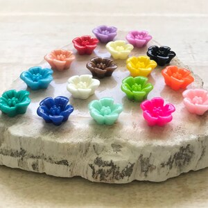 May include: Assortment of 16 colorful flower-shaped resin cabochons. The cabochons are arranged in a circle on a light gray stone surface. The colors include red, orange, yellow, green, blue, purple, pink, and white.