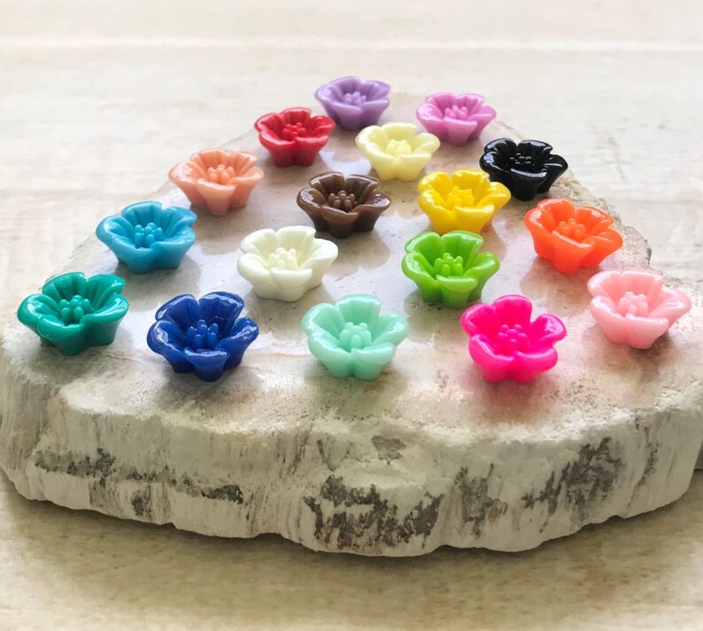 May include: Assortment of 16 colorful flower-shaped resin cabochons. The cabochons are arranged in a circle on a light gray stone surface. The colors include red, orange, yellow, green, blue, purple, pink, and white.