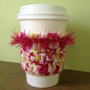 May include: A white coffee cup with a crocheted pink, white, and green cozy. The cozy is decorated with a pink and silver tinsel trim.