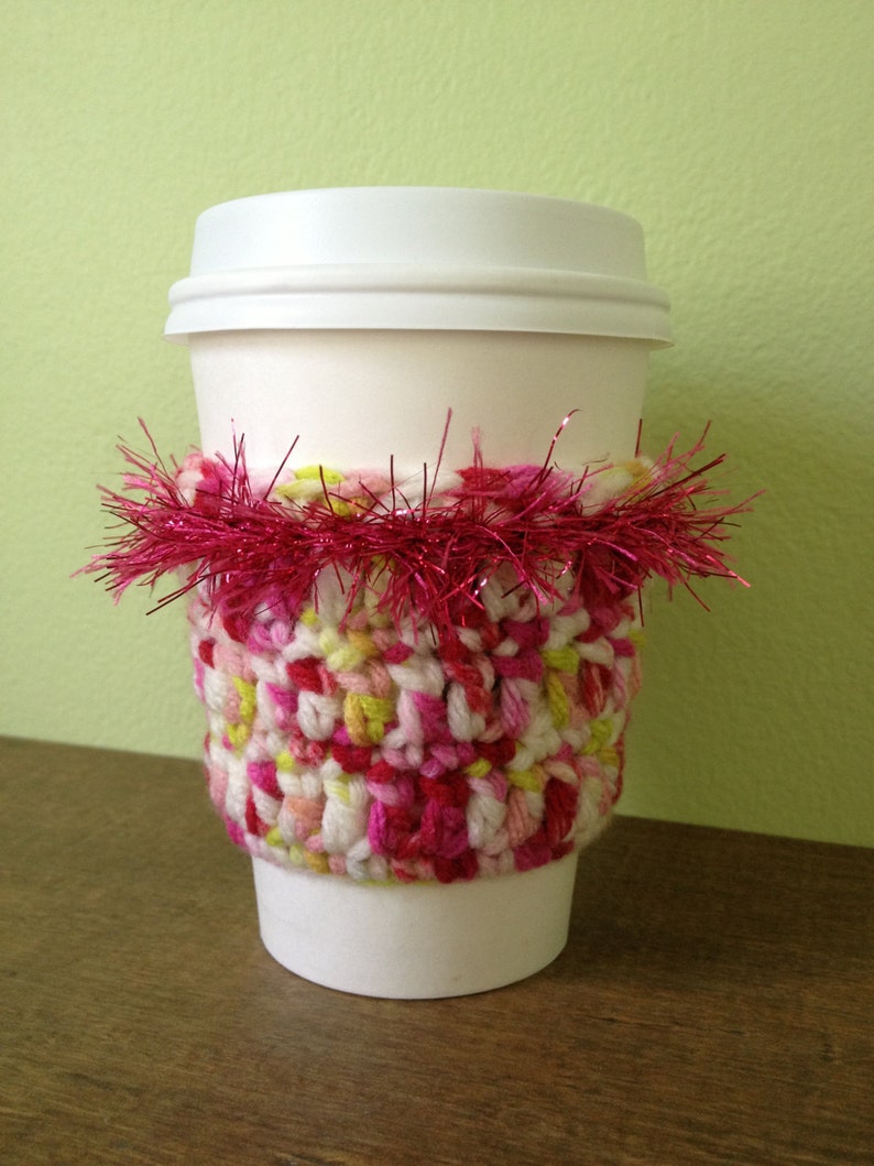 May include: A white coffee cup with a crocheted pink, white, and green cozy. The cozy is decorated with a pink and silver tinsel trim.