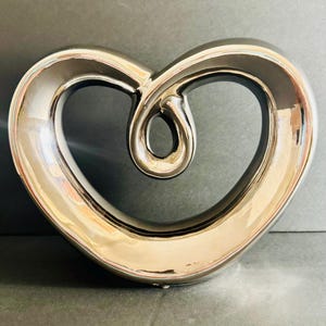 May include: A metallic silver, heart-shaped decorative sculpture with a smooth, reflective surface. The object features a continuous, curved form that loops inward at the top center, creating an open, hollow space within the heart shape.