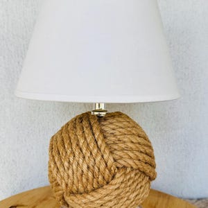 May include: A table lamp featuring a spherical base wrapped in thick, natural-coloured jute rope. The lamp is topped with a simple, solid white conical lampshade and rests on a clear, circular acrylic base that provides a stable foundation.