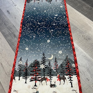 May include: A quilted table runner with a winter scene. The design features a snowy landscape with deer, pine trees, and a crescent moon. The edges are trimmed with red plaid fabric. The color palette includes shades of blue, white, and red.