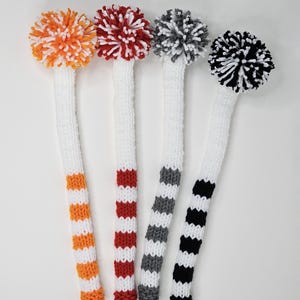 Puede incluir: Cuatro fundas para la cabeza de los palos de golf con pompones. Las fundas son blancas con rayas de color naranja, rojo, gris y negro.