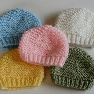 May include: Five knitted hats in different colors: light blue, pink, yellow, green, and white. The hats are all made with a simple stitch pattern and have a rounded crown.
