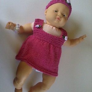 May include: A pink knitted dress with white trim and floral accents, worn by a doll with a pink knitted hat with white flowers.