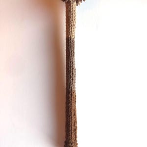 May include: A handmade, brown and tan knitted bottle stopper. The stopper features a pom-pom top and a long, slender stem. The color gradient transitions from light tan to dark brown. The stopper is designed to fit into a bottle.